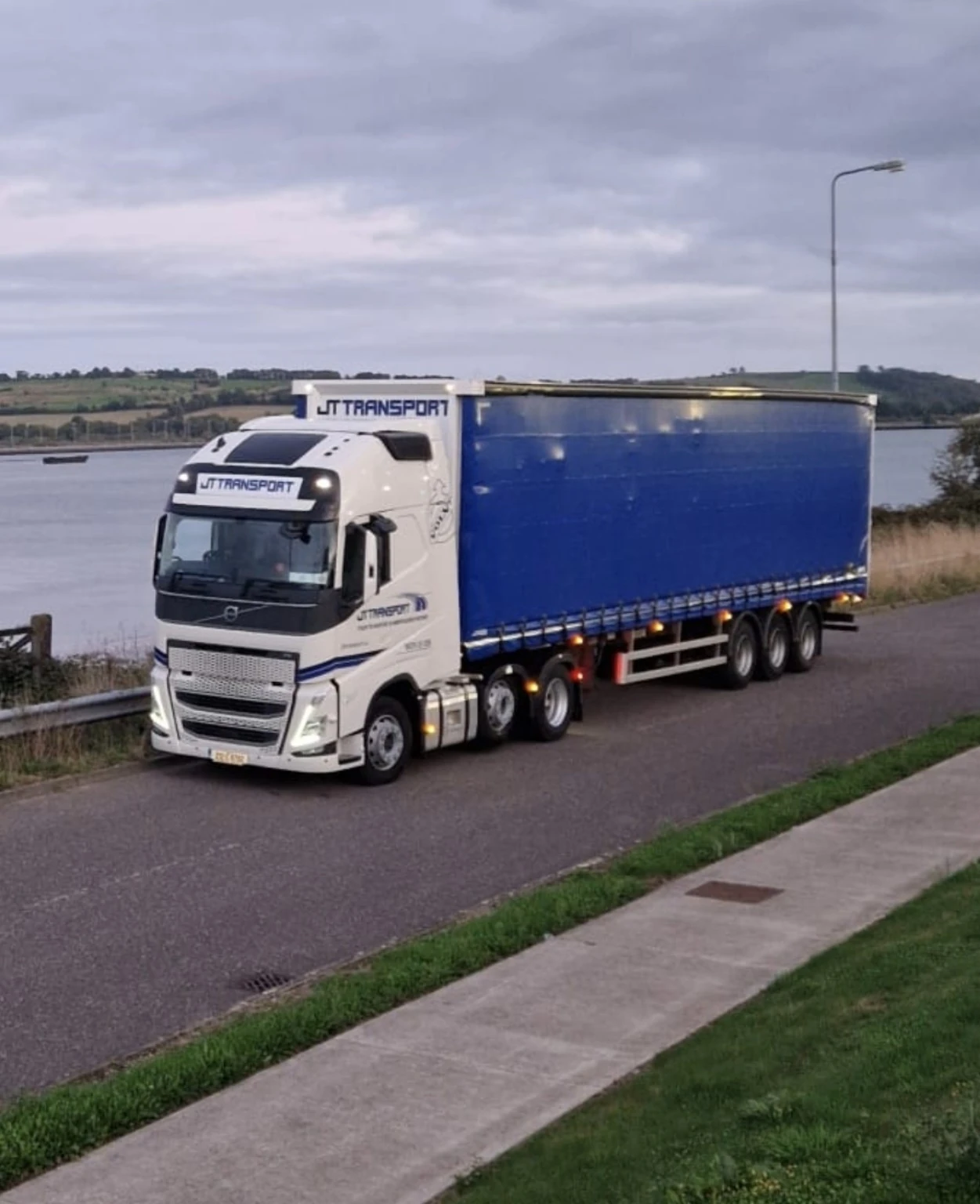 JT-branded Volvo articulated truck with a blue curtainsider trailer on a road beside the water.